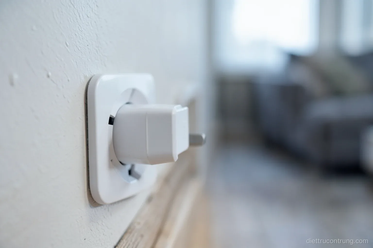 Macro shot of a small white plug-in device in a wall socket beside a wooden skirting board, soft bokeh of a living room behind, cool neutral light, no visible markings