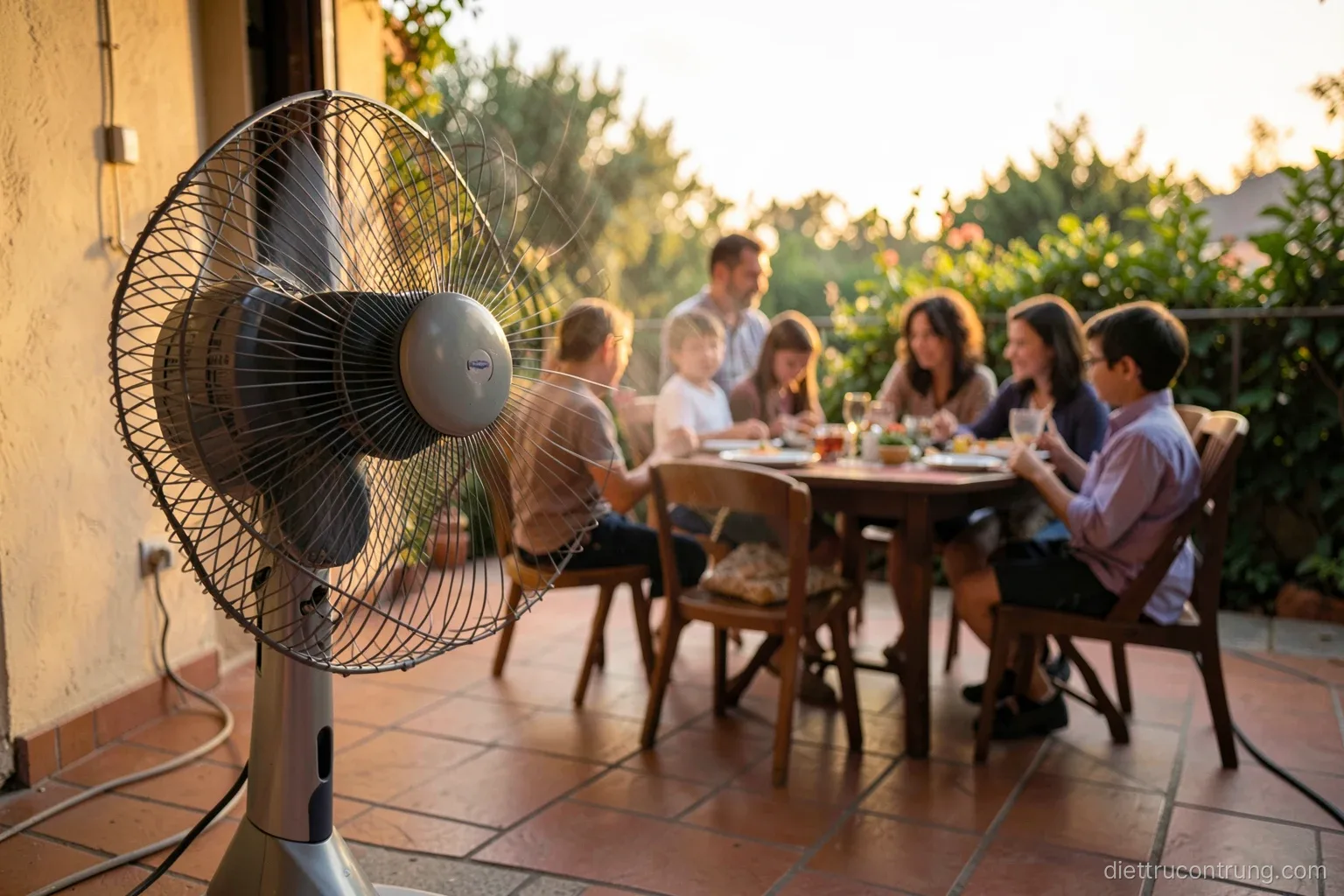 Low angle view of a standing fan on a tiled veranda blowing toward a small family dinner table, people seen from behind, warm evening light, gentle motion blur on blades